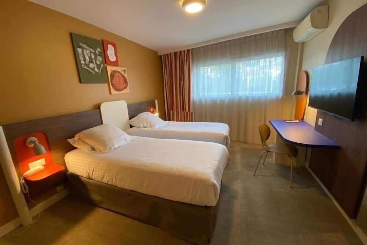 Habitación de hotel con dos camas, un escritorio, una silla, televisión y cuadros en la pared.