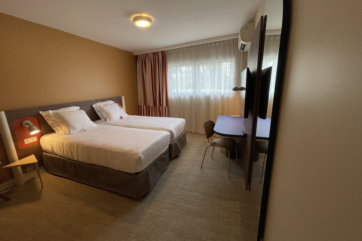 Habitación de hotel con dos camas, un escritorio, una silla, un televisor y una ventana con cortinas.