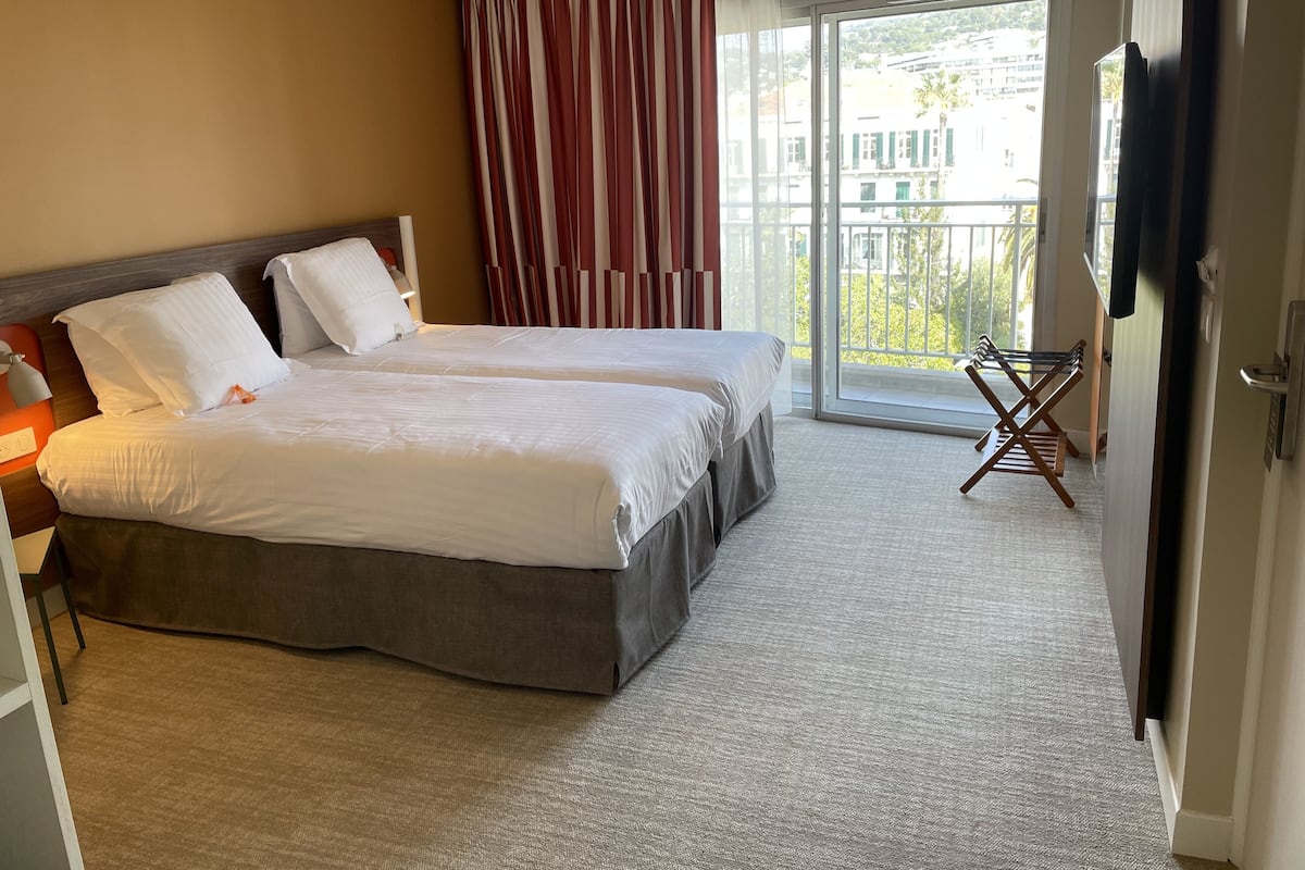 Habitación de hotel con dos camas, un balcón con una silla y un televisor en la pared.
