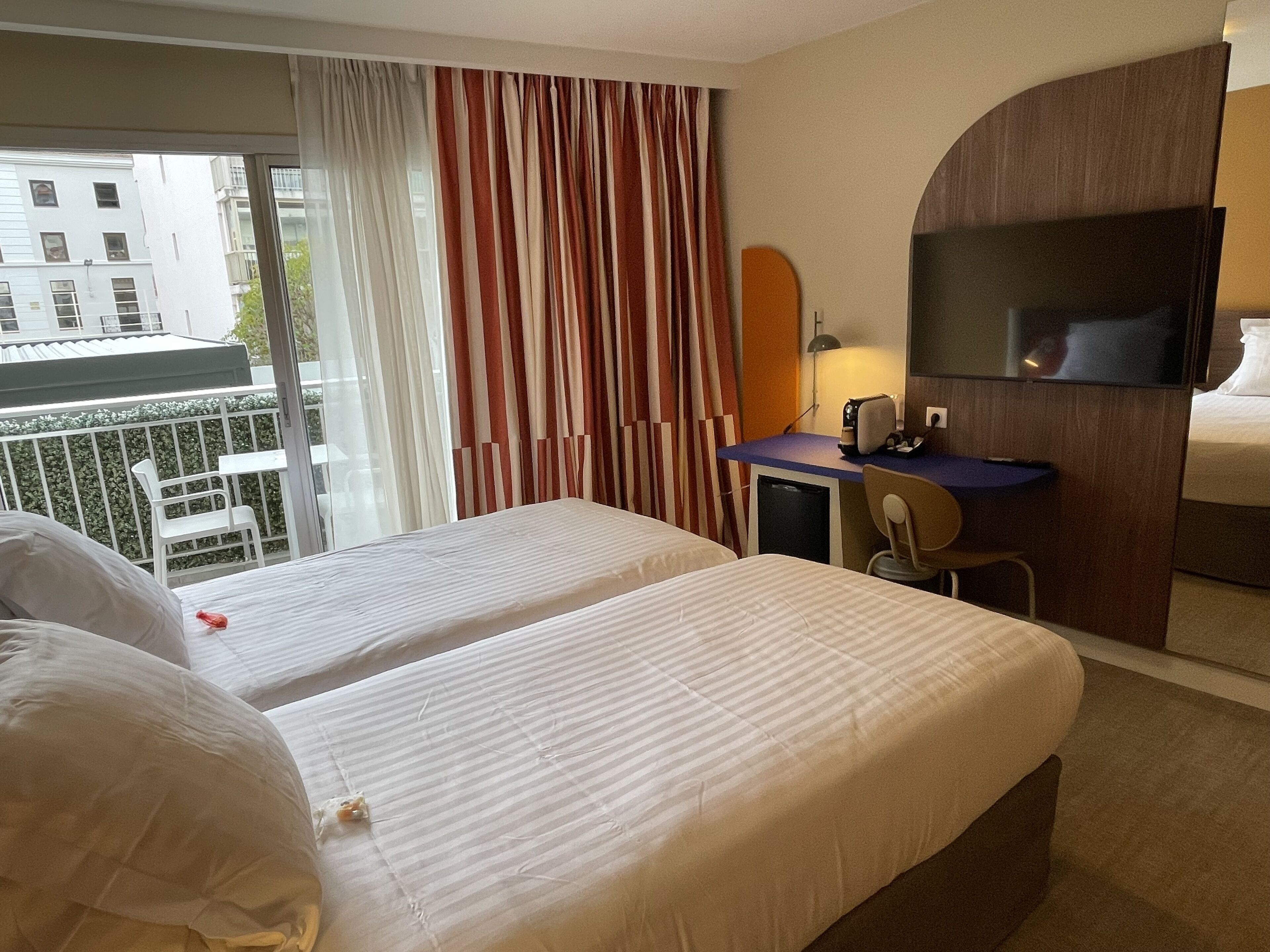 Habitació d'hotel amb dos llits, una taula de treball, una cadira, un televisor i un balcó amb cortines.