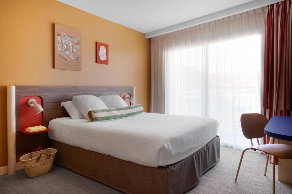 Una habitación de hotel con una cama grande, una mesita de noche con lámpara, una silla y una ventana con cortinas.