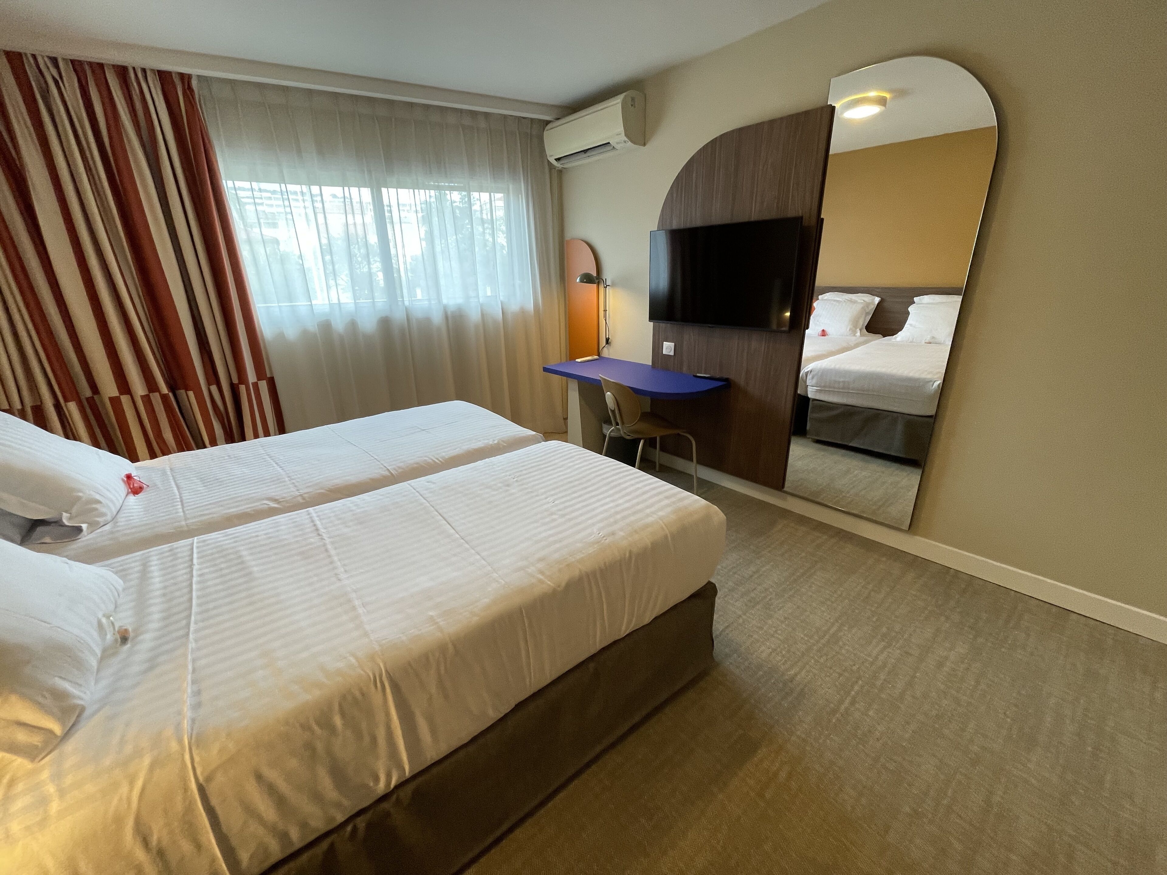 Une chambre d’hôtel avec deux lits, un bureau, une chaise, une télévision et un grand miroir.