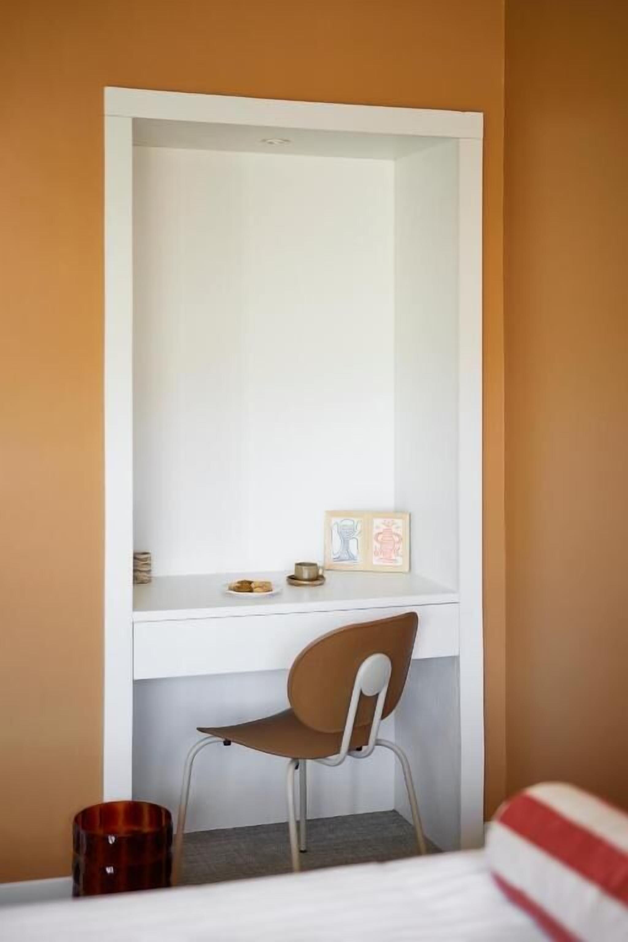 A compact room with a white desk, a brown chair, a small shelf with a picture frame and a cup, and a red and white striped pillow on the bed.