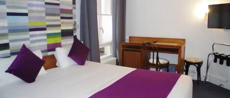 A hotel room with a large bed, a desk, and a chair. There is a television mounted on the wall and a window with curtains.