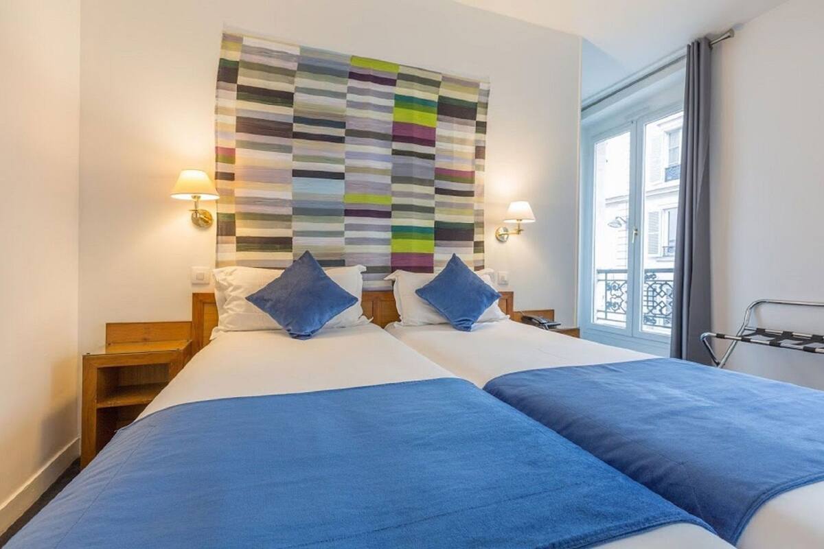A double bed with blue bedding, a colorful striped wall hanging, and a window with a view.