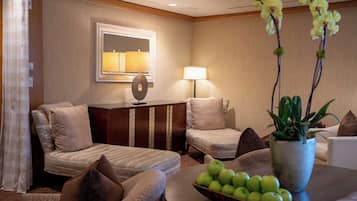 A hotel room with a bed, two armchairs, a desk with a lamp, a mirror, and a table with a bowl of apples and a potted plant.