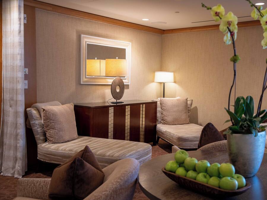 A hotel room with a bed, two armchairs, a desk with a lamp, a mirror, and a table with a bowl of apples and a potted plant.