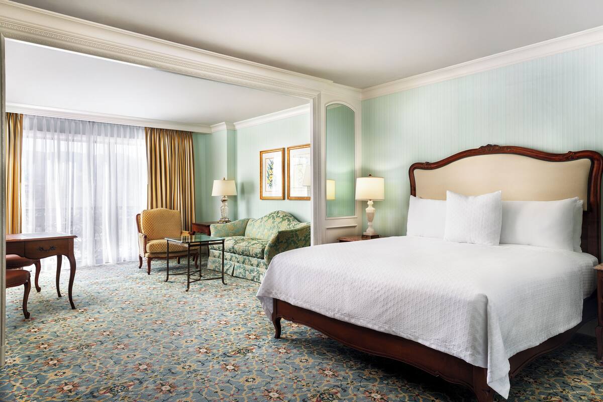 Ein Hotelzimmer mit einem großen Bett, einem Sessel, einem Schreibtisch und einem Fenster mit Vorhängen.