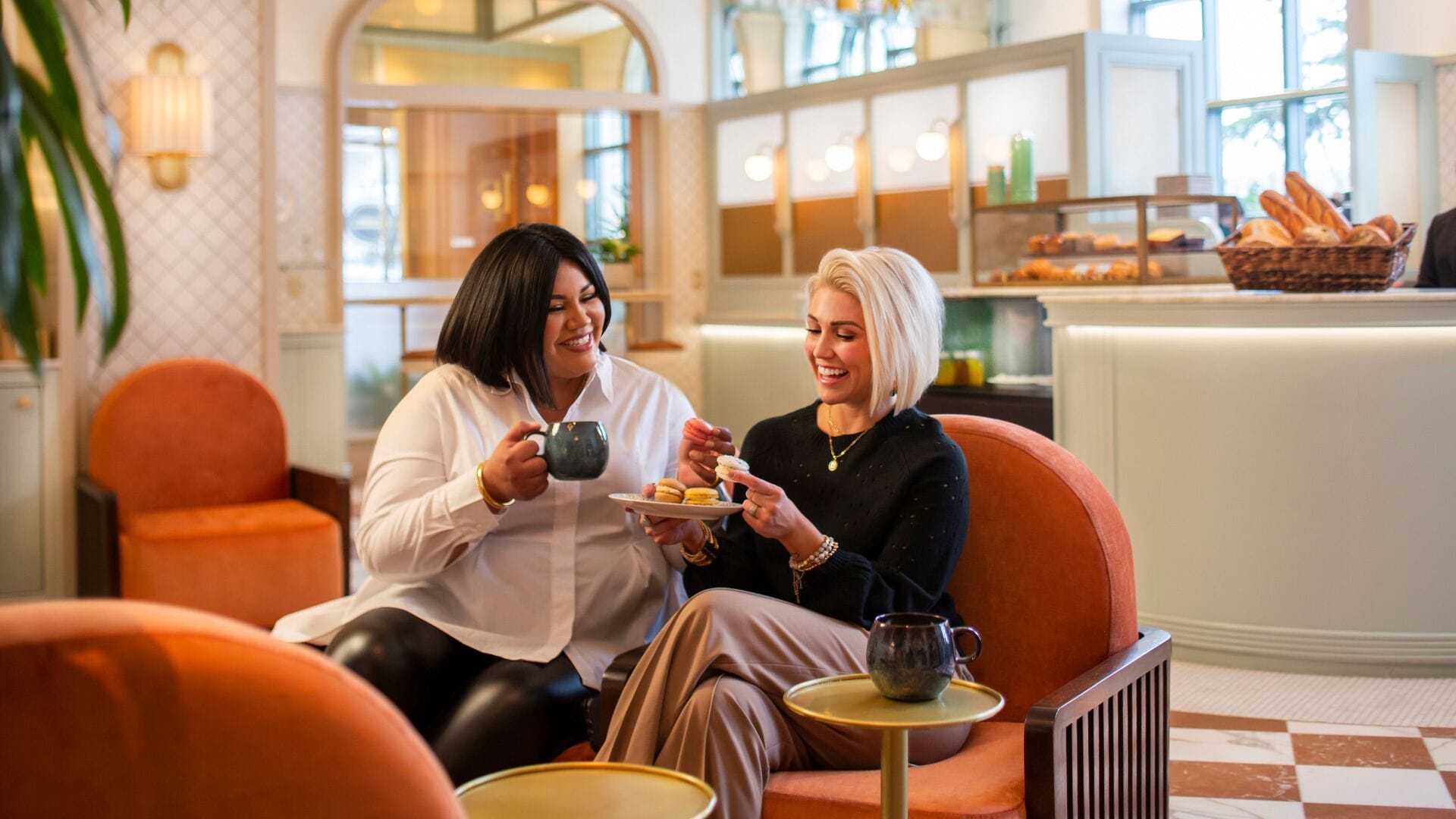 Zwei Personen sitzen auf orangefarbenen Stühlen in einem Café, in dem eine Theke mit Gebäck angeboten wird.