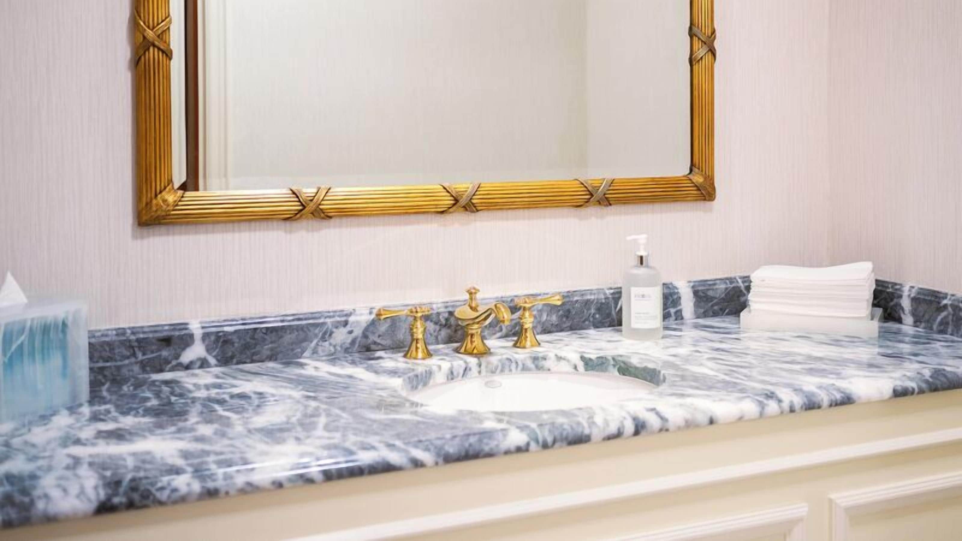 A bathroom with a marble sink, gold faucets, a large mirror with a gold frame, and a bottle of hand soap.