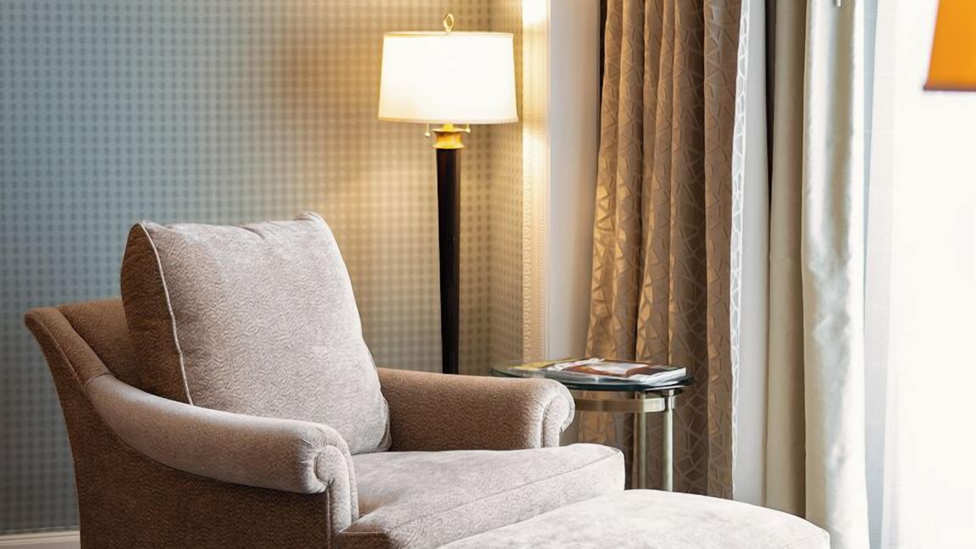 A plush armchair with a cushion, a small table with a magazine, a lamp, and patterned curtains.