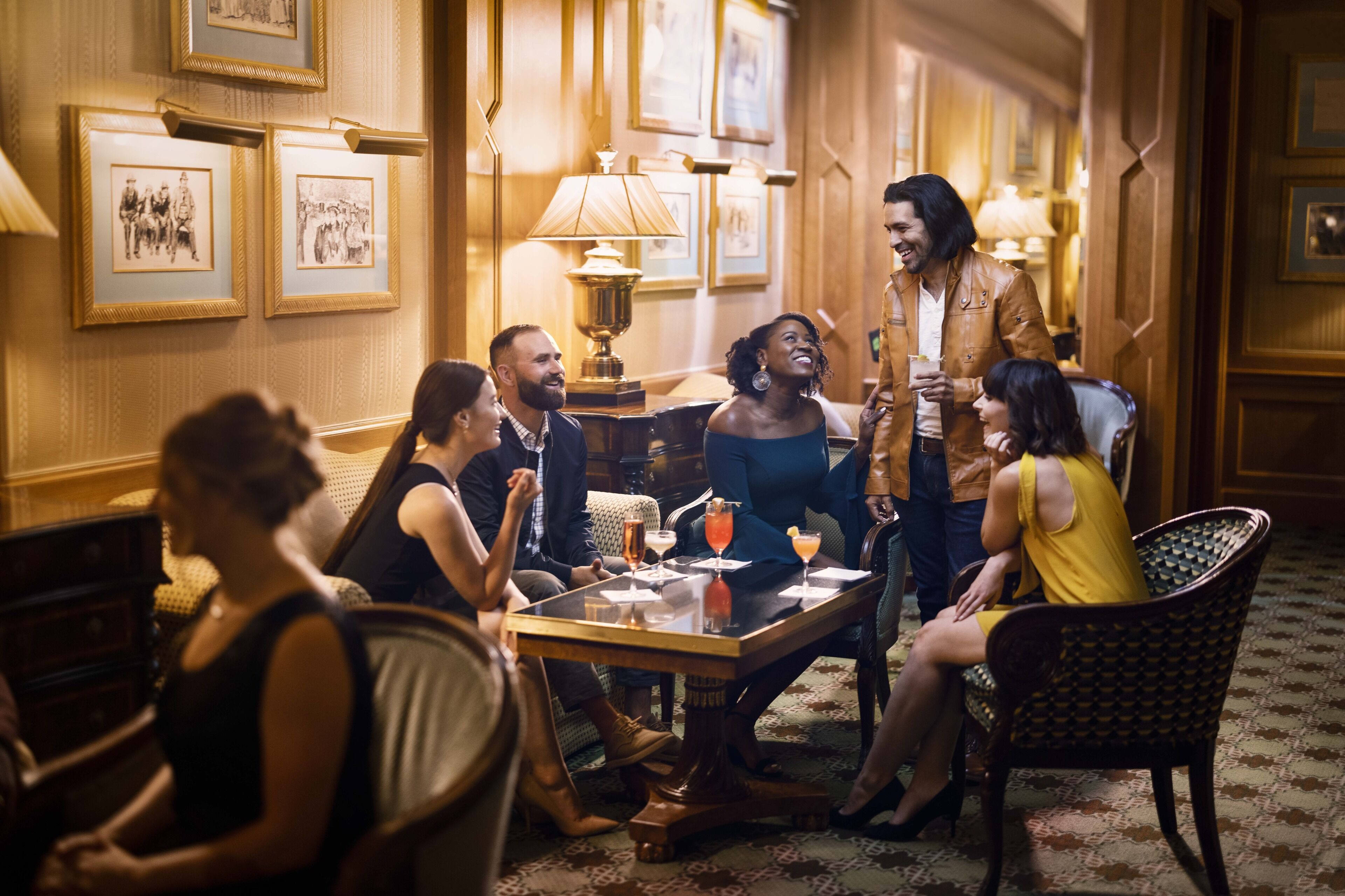 Un groupe de personnes est assis autour d’une table dans une pièce chaleureuse, éclairée d’une lumière douce, et les murs sont ornés de tableaux encadrés.