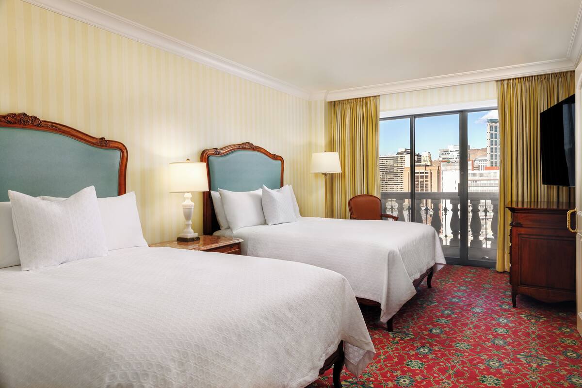 Ein Hotelzimmer mit zwei Betten, einem großen Fenster mit Blick auf die Stadt, einem Fernseher und einem Holzkasten.