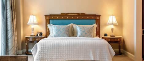 Una cama bien hecha con una colcha blanca acolchada, flanqueada por dos mesitas de noche con lámparas, un cabecero de madera y vistas a una puerta.