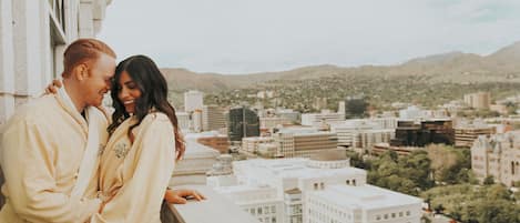 Una pareja parada en un balcón con vista a un paisaje urbano que incluye edificios y colinas al fondo.