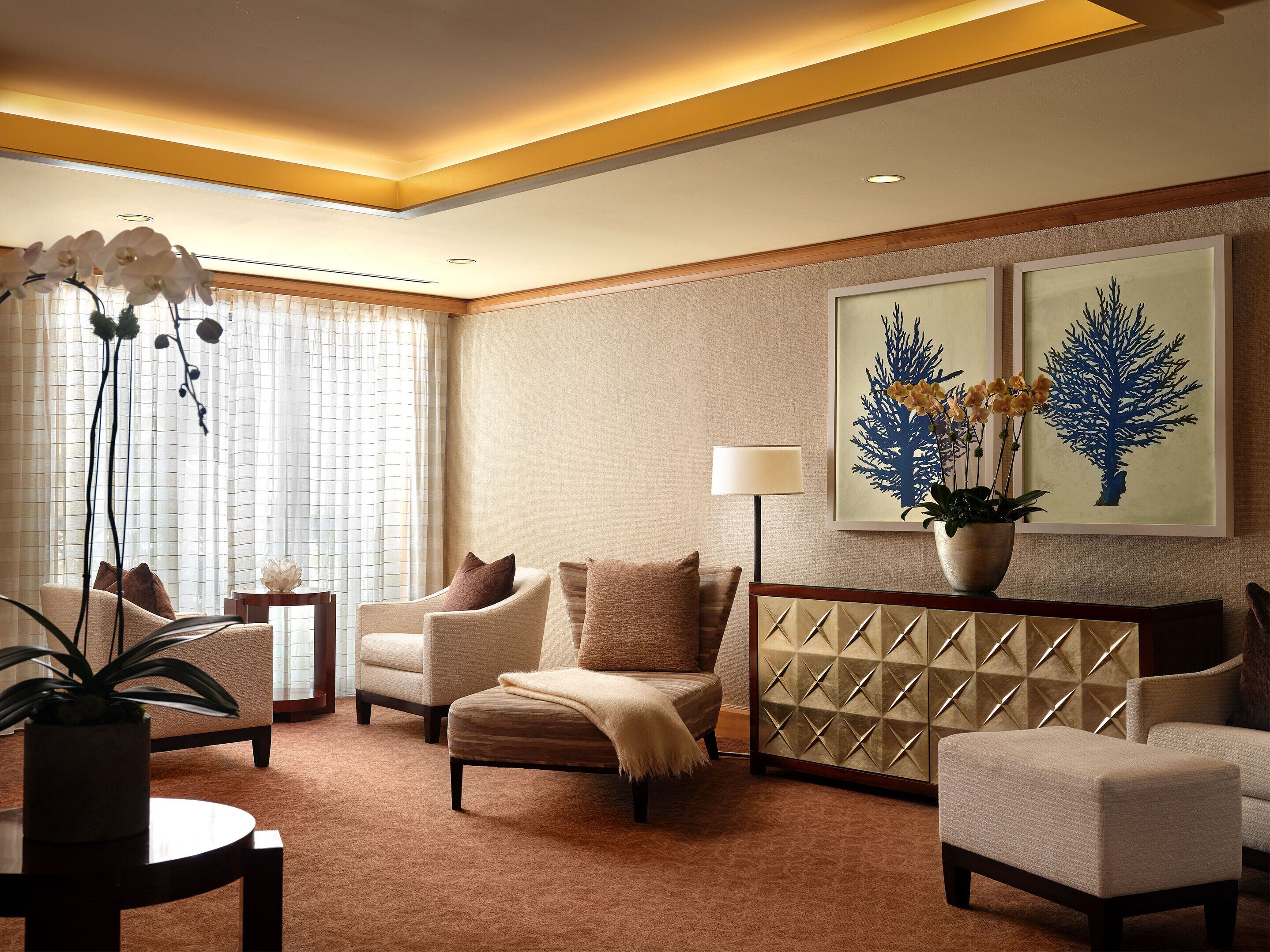 A well-lit living room with a sofa, armchairs, a coffee table, and a decorative plant. There are two framed artworks on the wall and a lamp on a side table.