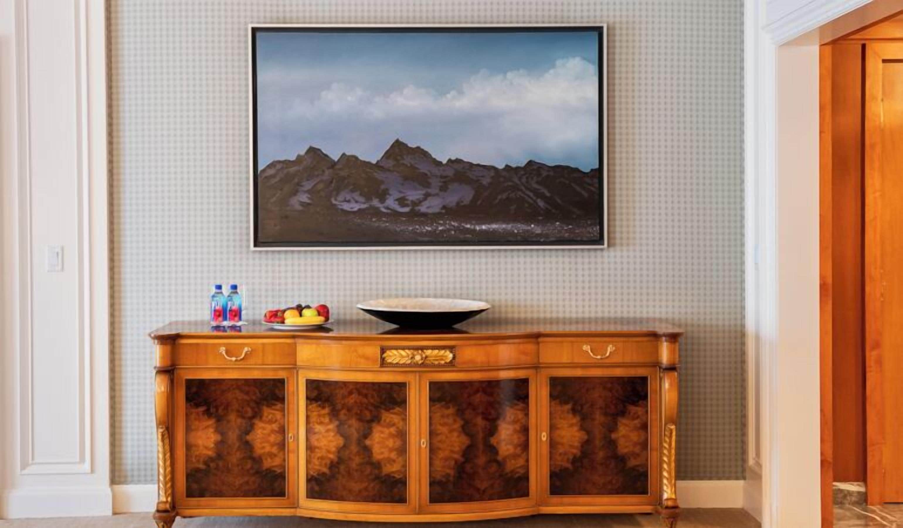 A wooden console table with a bowl and fruit, a flat-screen TV above it, and a patterned wall.