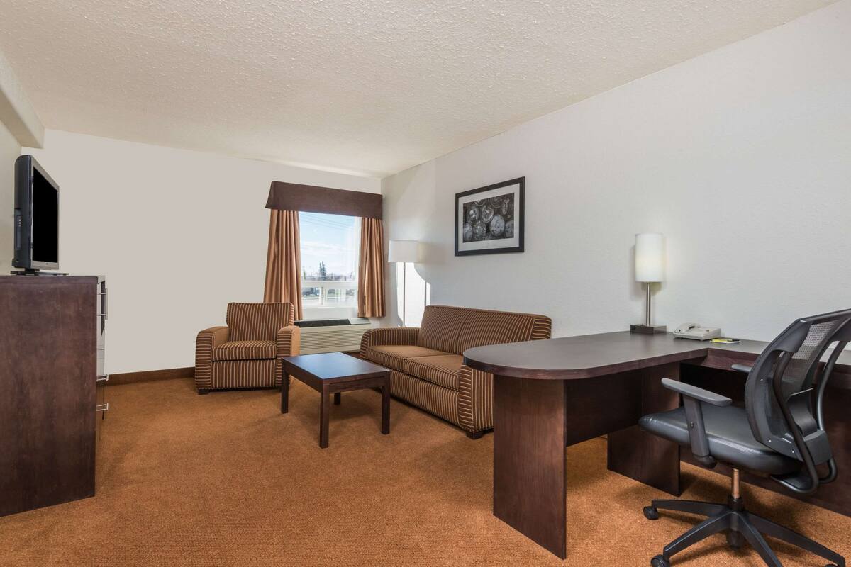 A hotel room with a brown carpet, a sofa, a chair, a desk, a computer, and a television.
