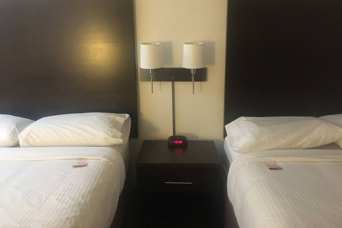 A hotel room with two beds, each with white linens and pillows, a nightstand with a clock, and two wall-mounted lamps.