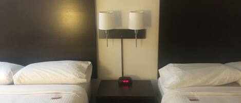 Quarto de hotel com duas camas, cada uma com roupa de cama e almofadas brancas, uma mesinha de cabeceira com relógio e duas luminárias fixadas na parede.