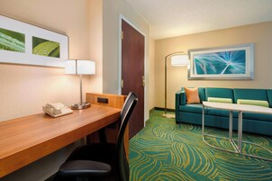A hotel room with a wooden desk, a telephone, a lamp, a green patterned carpet, a teal sofa, and a framed abstract painting.