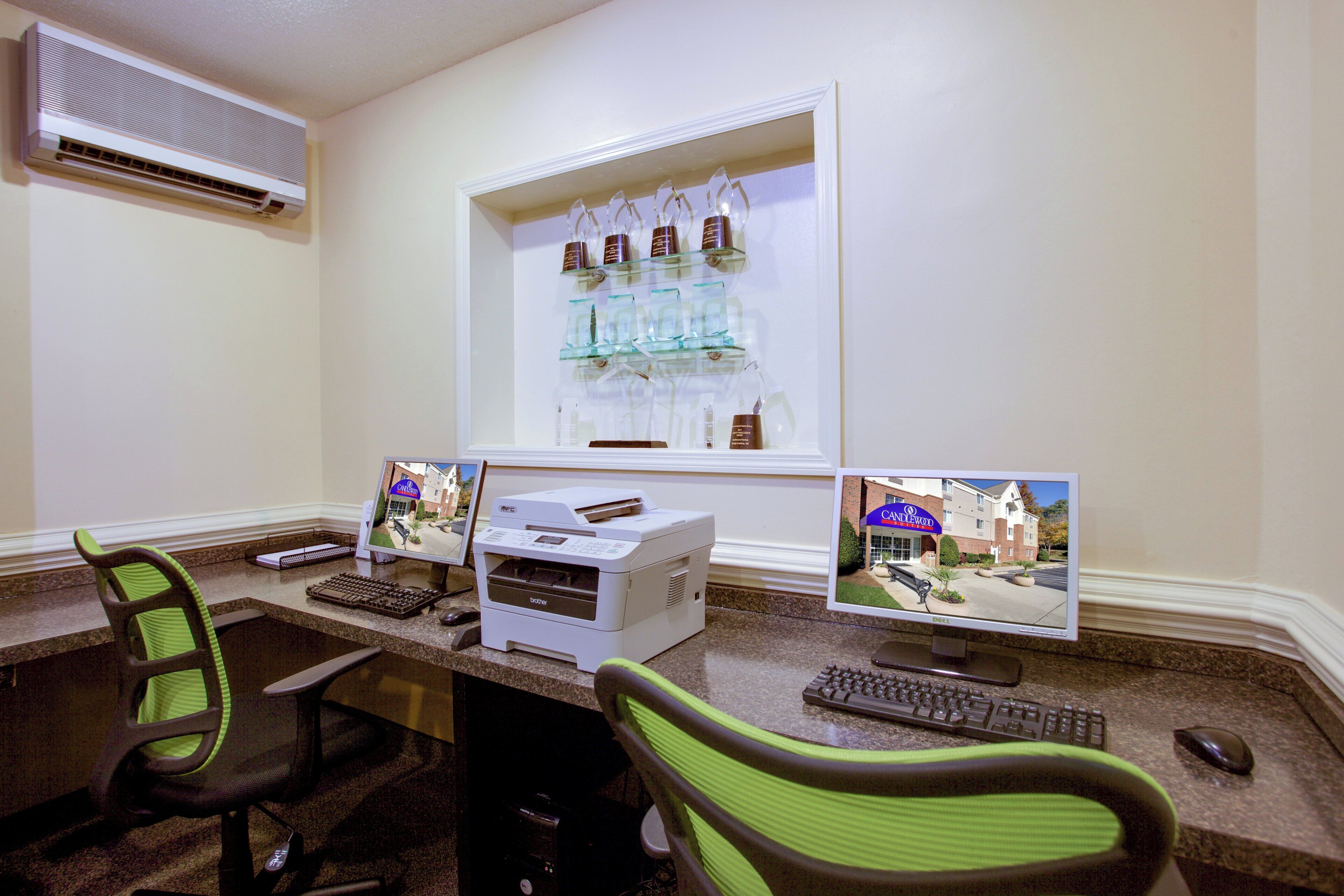 Habitación de hotel con un escritorio, dos monitores, una impresora, un teclado, una silla y una ventana que ofrece vistas del exterior del hotel.