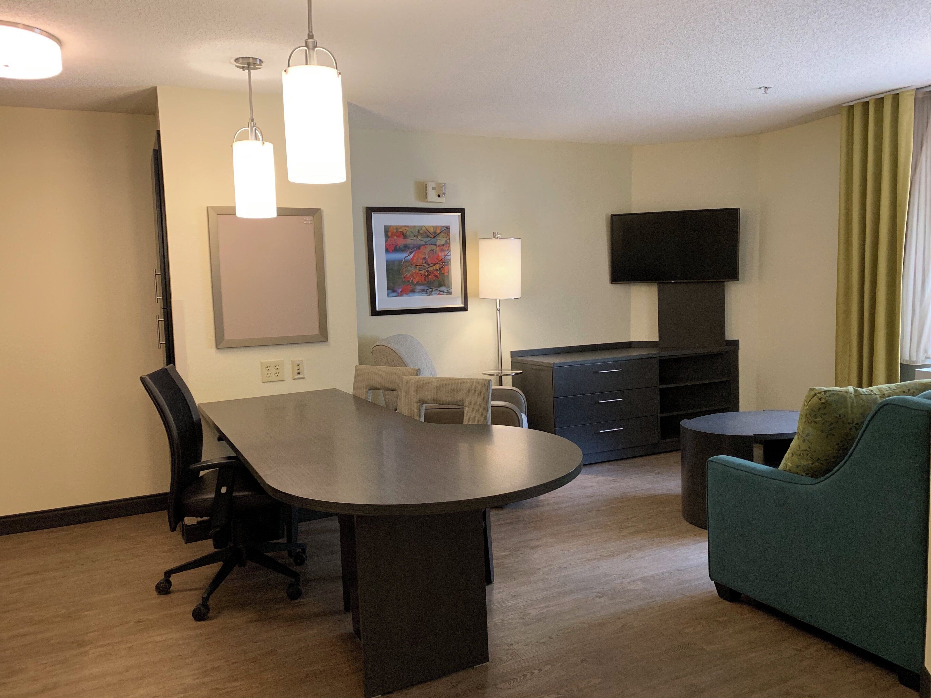 Ein Hotelzimmer mit Esstisch, Schreibtischstuhl, Sofa, Fernseher und Kunstwerk an der Wand.