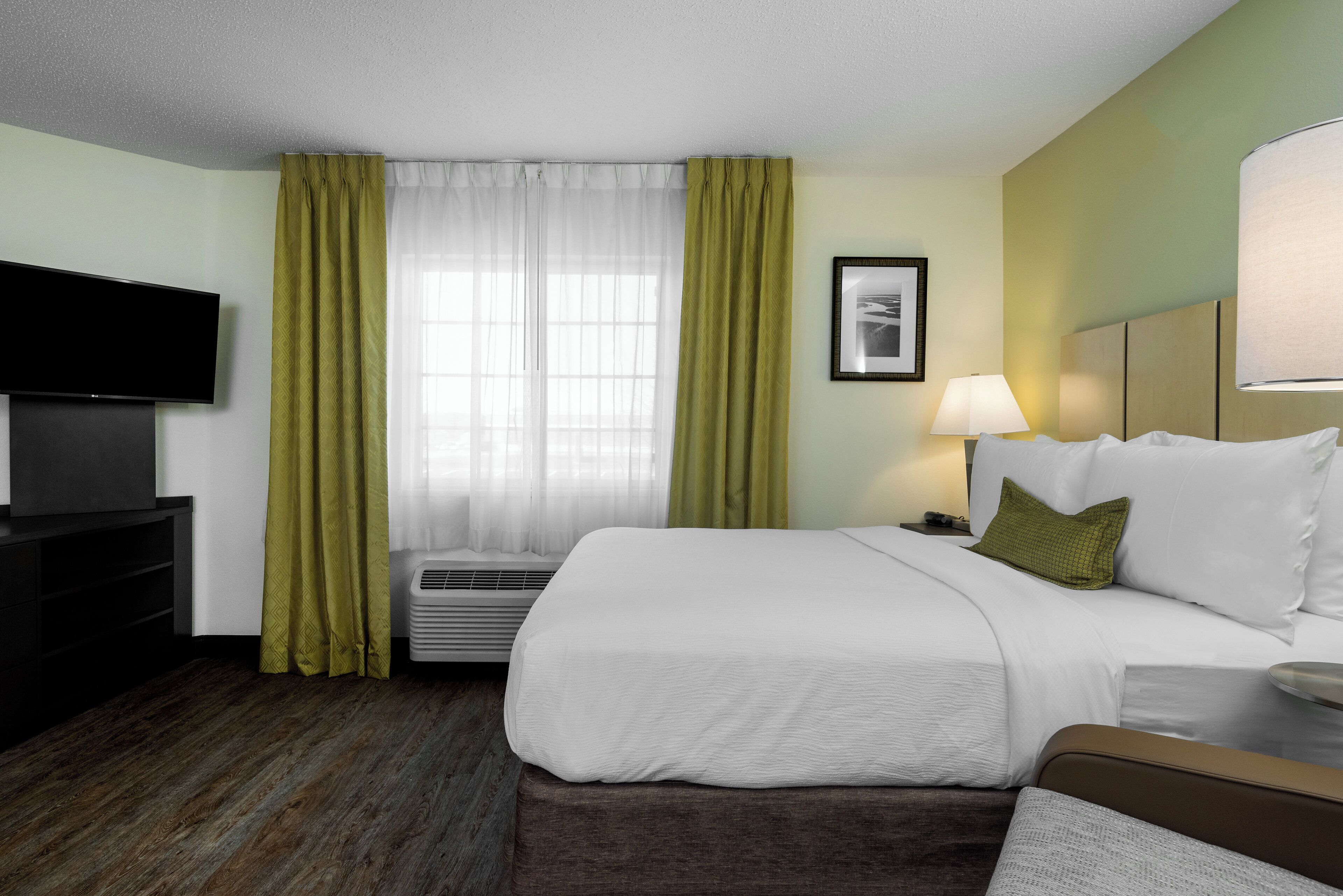Une chambre d’hôtel avec un lit, une télévision, une fenêtre avec des rideaux, un tableau au mur et une lampe de chevet.