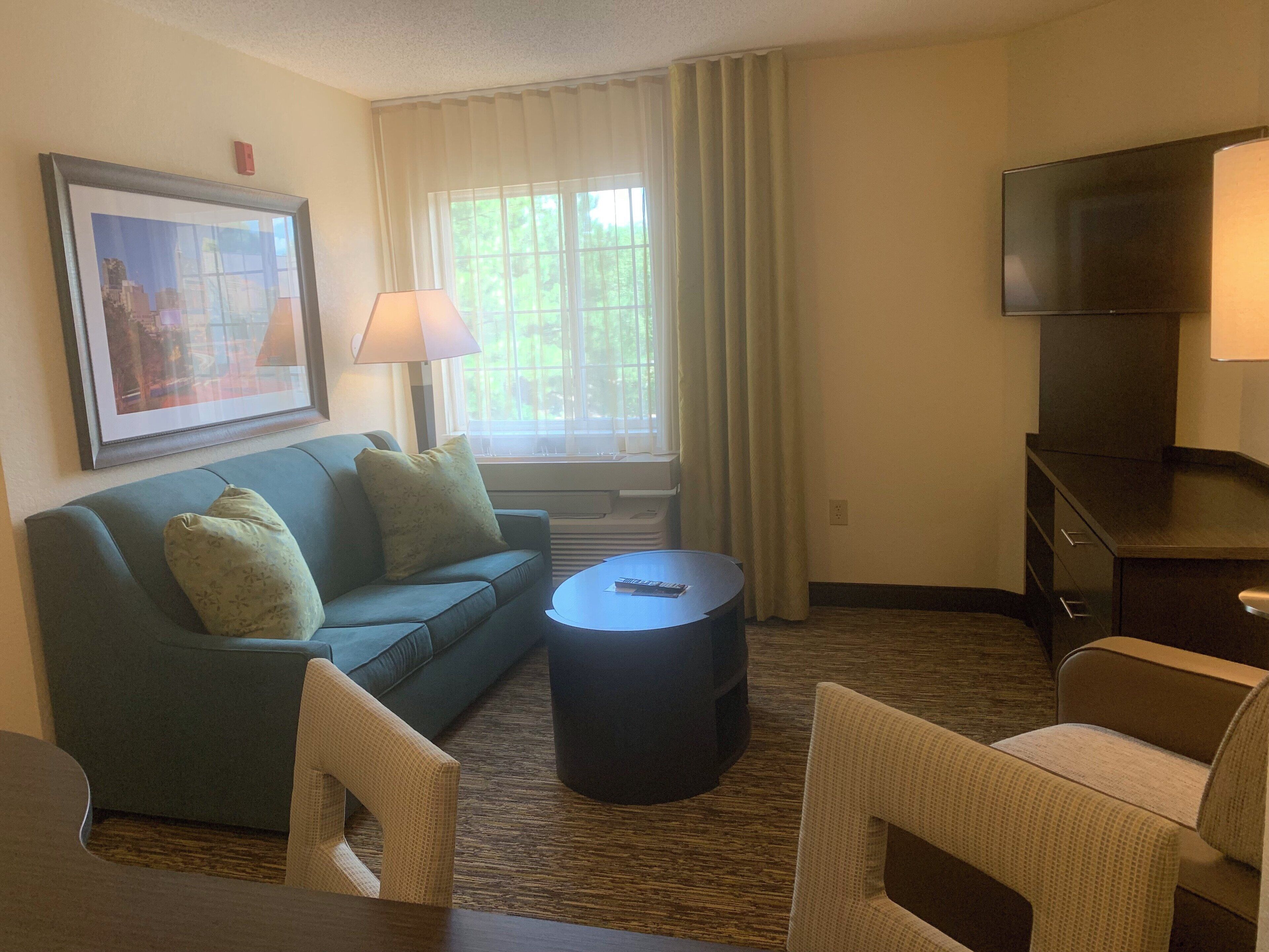 A hotel room with a sofa, a round coffee table, a TV, and a window with curtains.