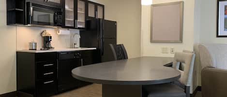 Een moderne keuken met zwarte kasten, een magnetron en een koffiezetapparaat. Er is een eettafel met stoelen en een bank in de woonkamer.