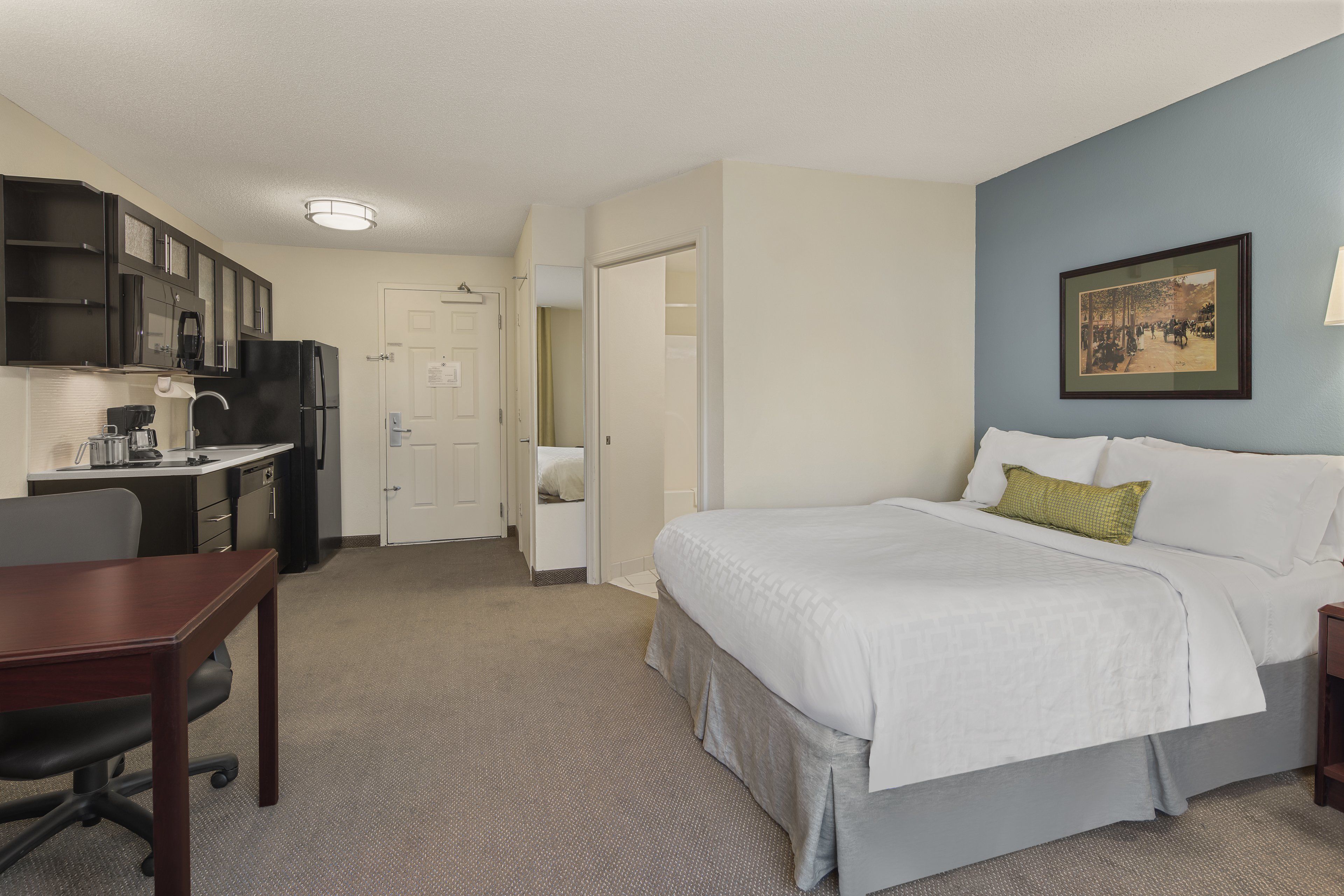 Una habitación de hotel con cama, escritorio, silla, kitchenette y puerta que da a un baño.