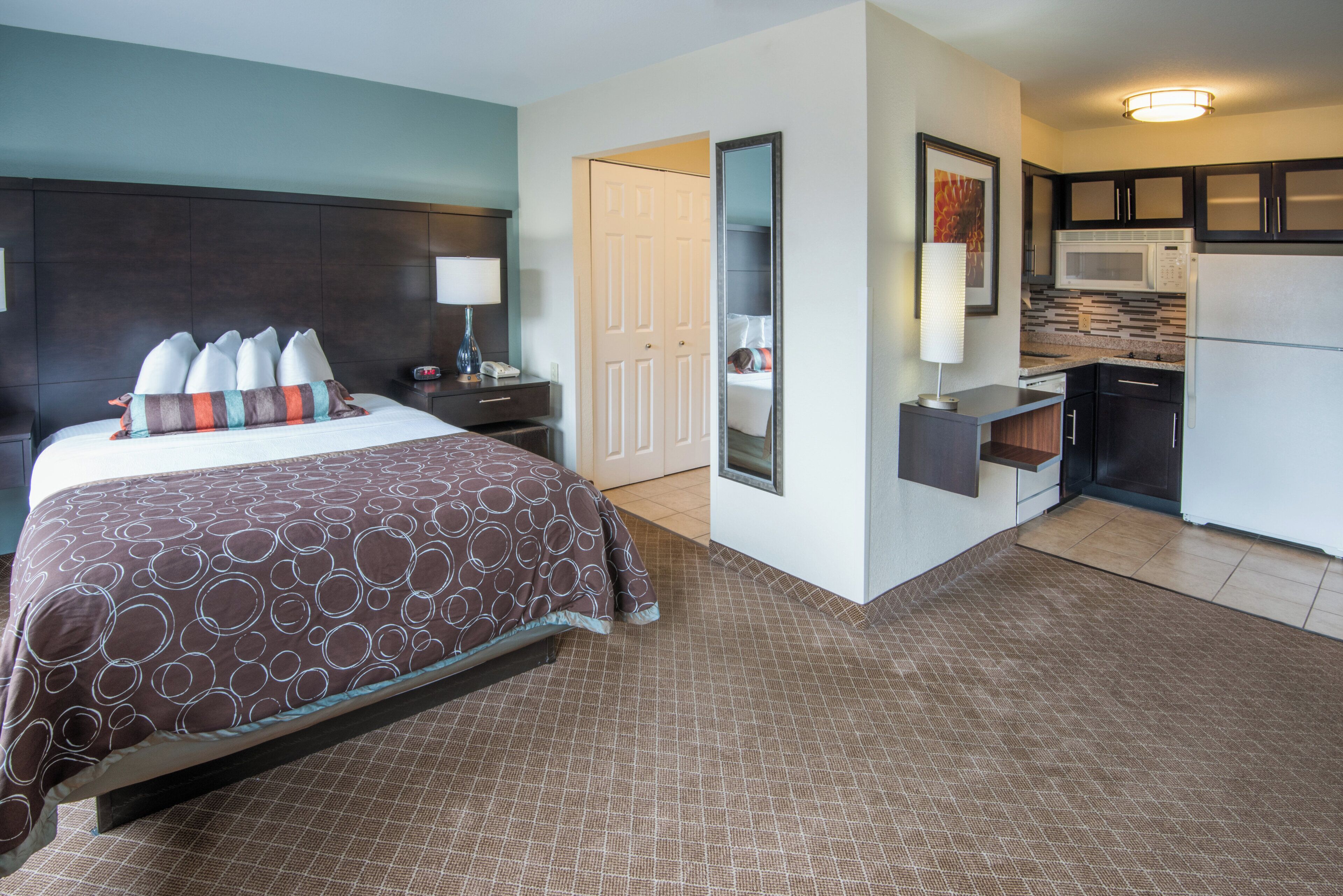 Habitación de hotel con cama, mesita de noche, lámpara y una zona de kitchenette con microondas y refrigerador.