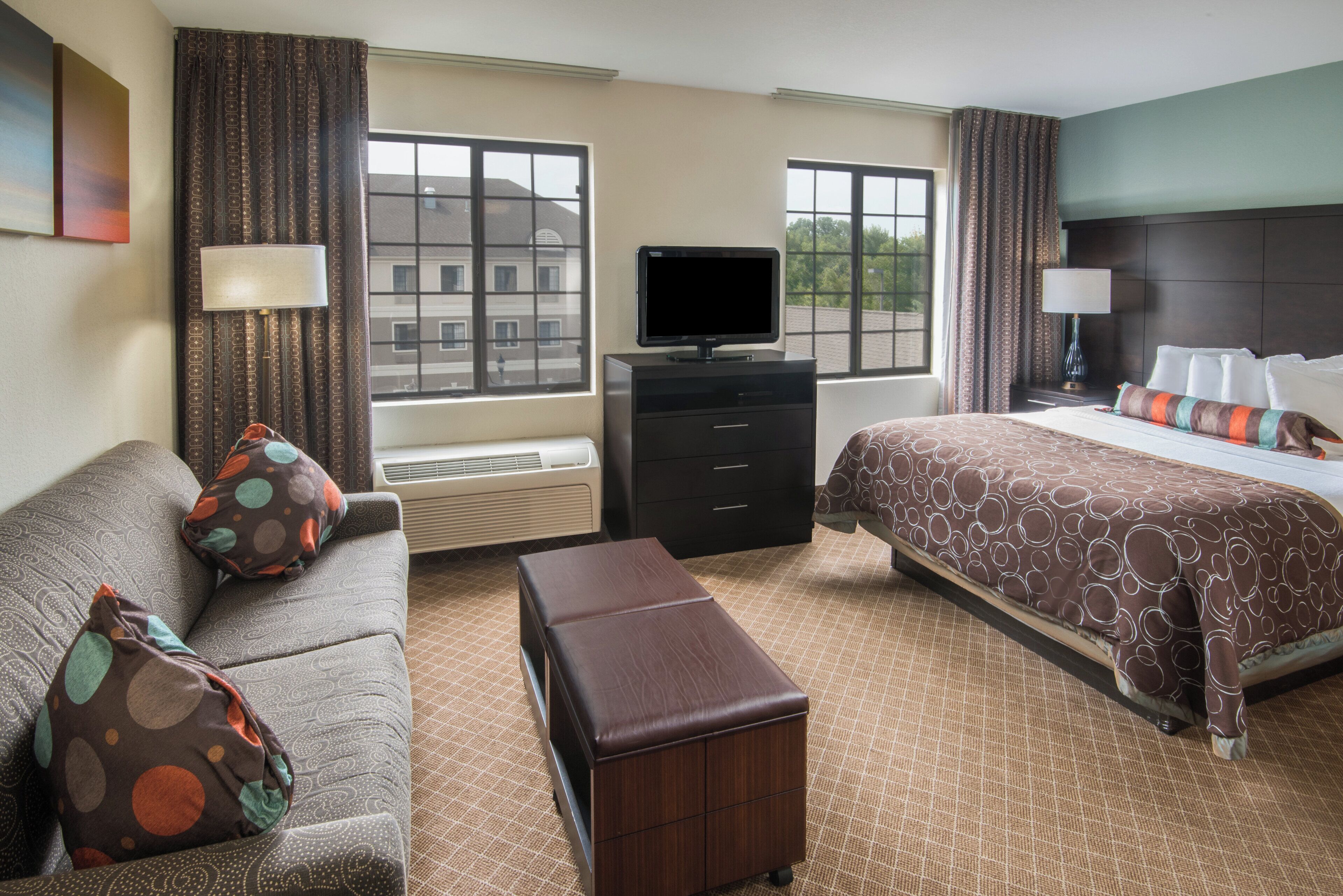 Habitación de hotel con cama, sofá, televisión, escritorio y ventana con vista a edificios.