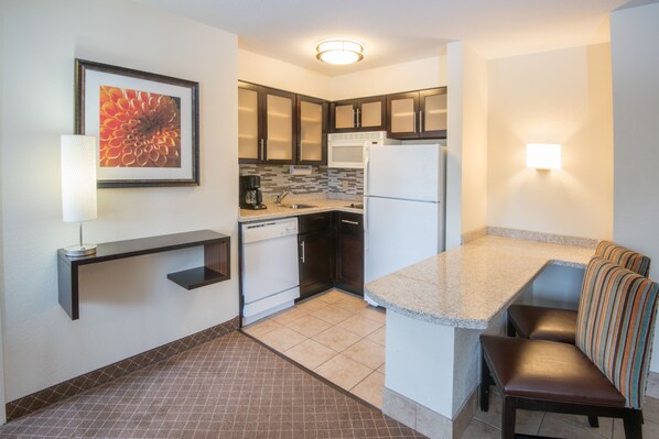 A compact kitchen with a microwave, refrigerator, and a coffee maker. There is a small seating area with a bench and a wall-mounted shelf.