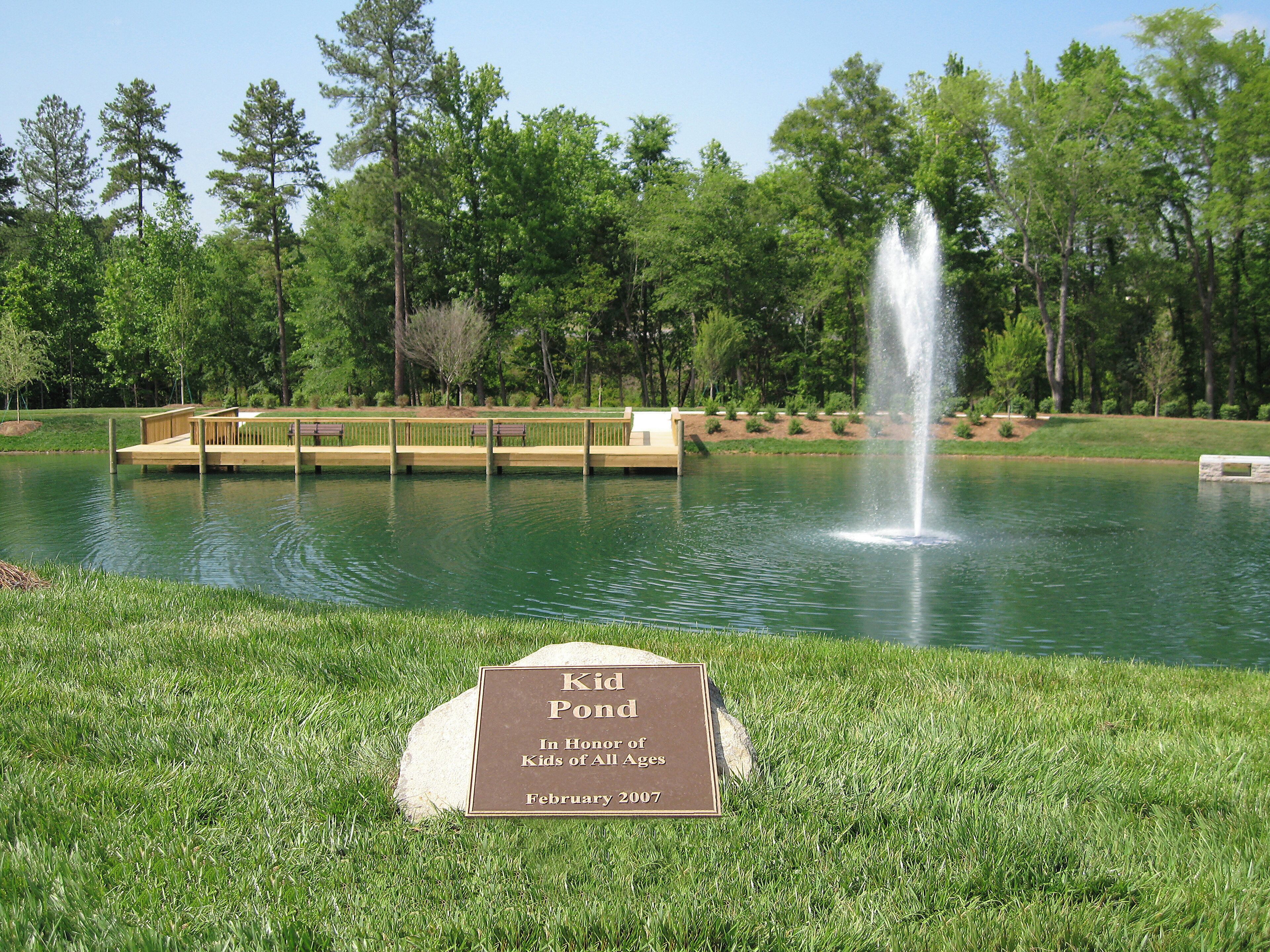 A park with a wooden dock over water, a fountain, and a plaque on the grass.