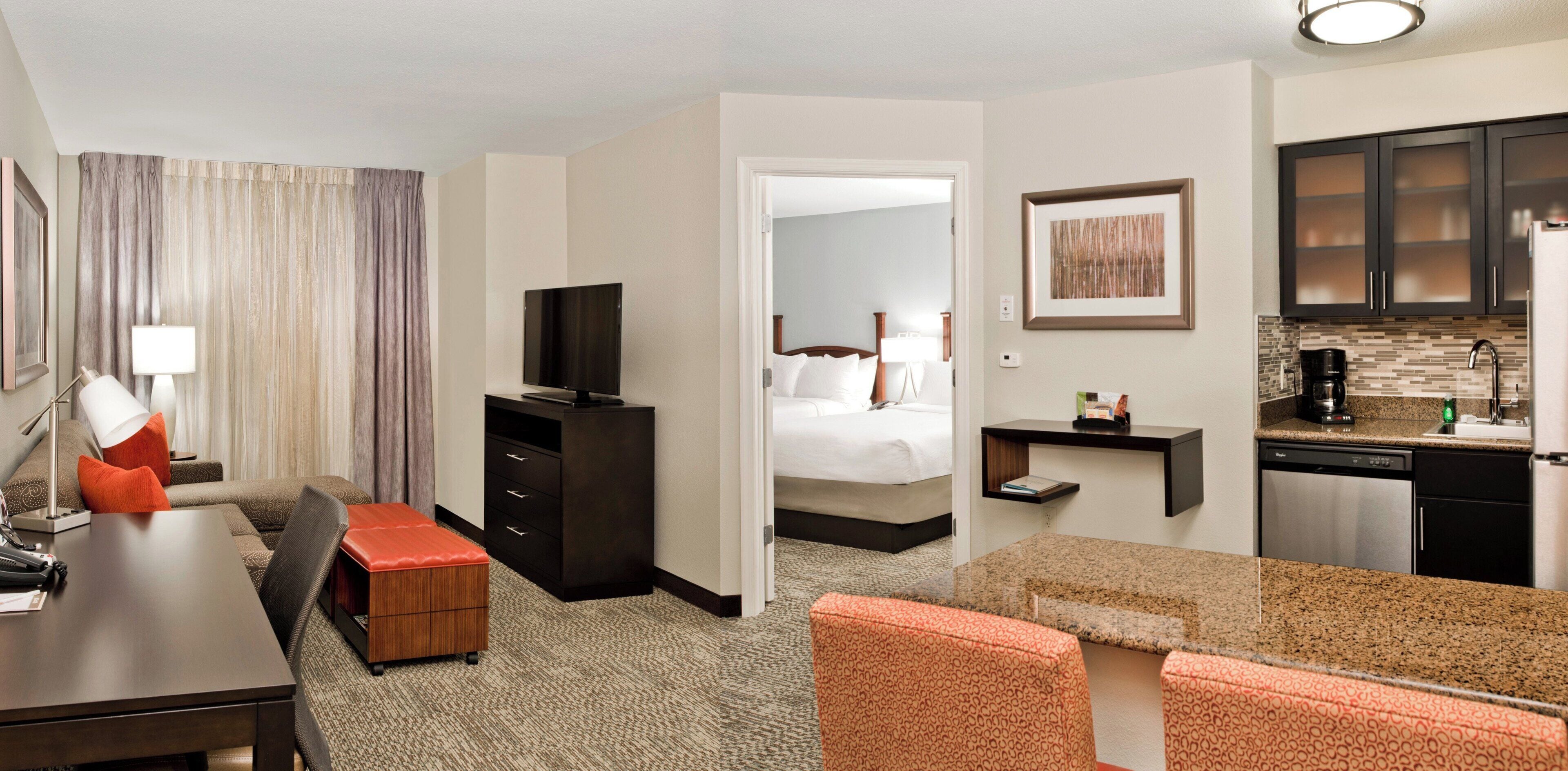 A hotel room with a desk, a television, a bed, a small kitchenette, and a bathroom.