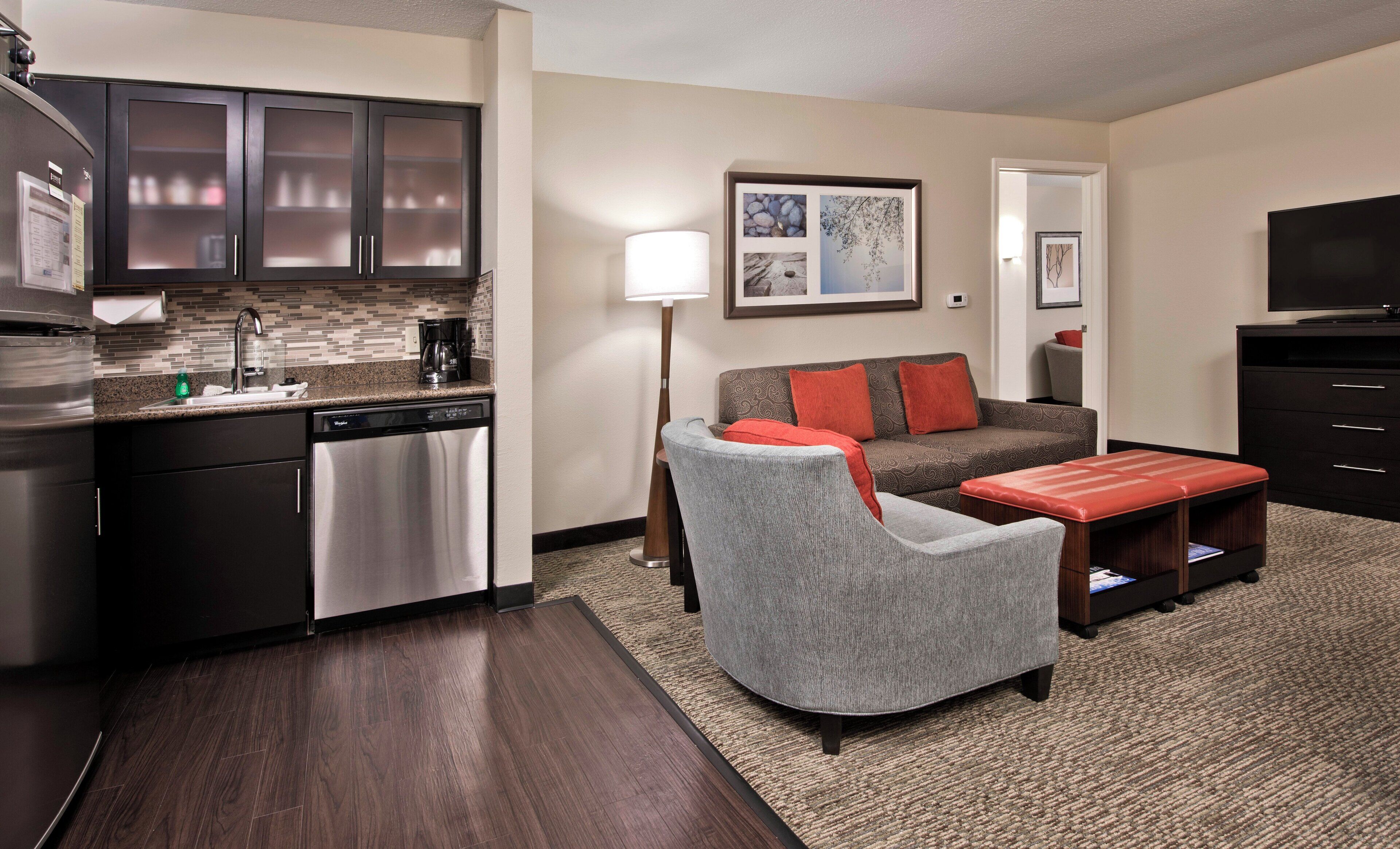 Una habitación de hotel moderna con zona de cocina, un sillón gris, un sofá con cojines rojos, un televisor de pantalla plana y una mesita.