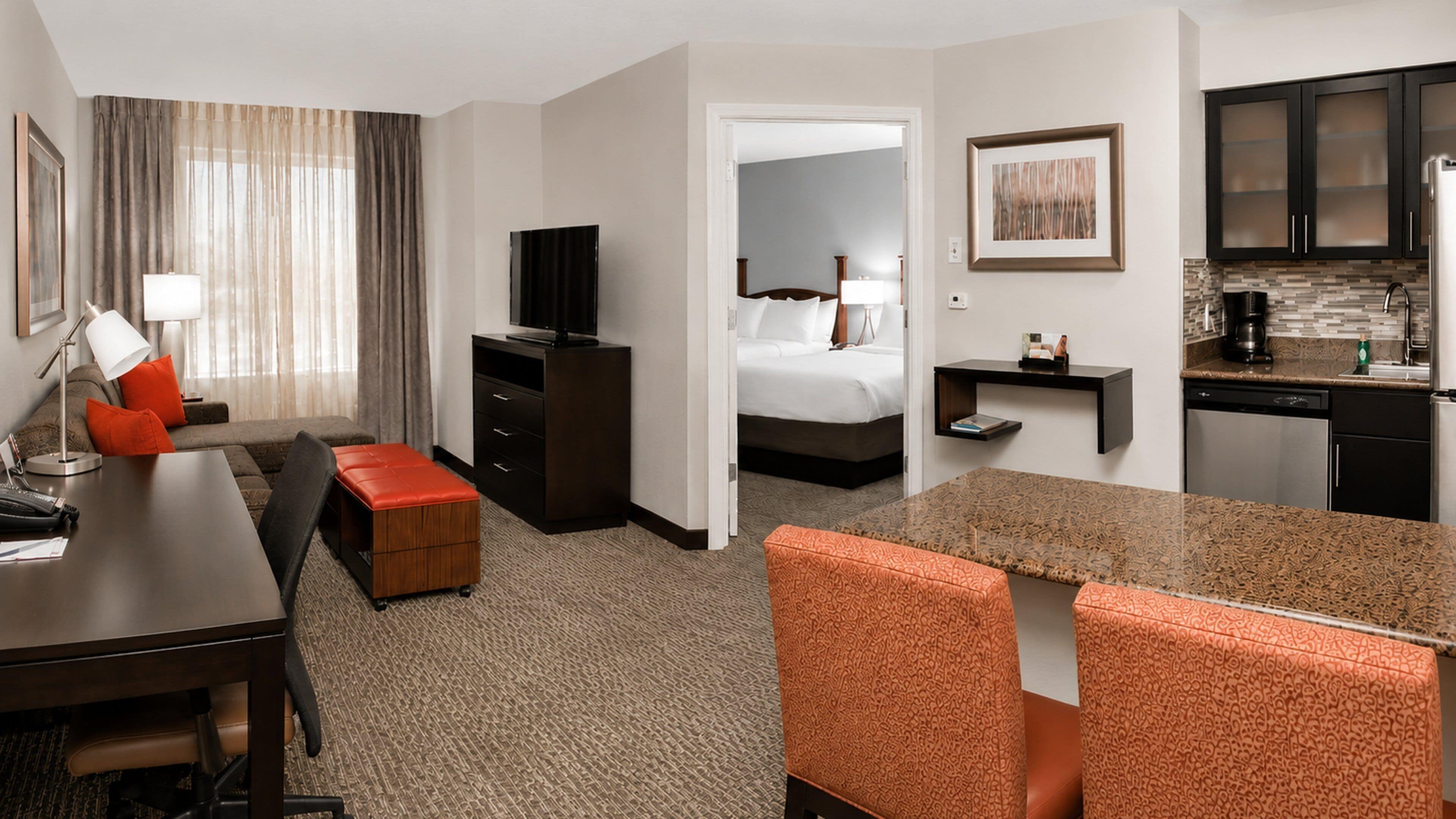 Une chambre d’hôtel comprenant un lit, un bureau avec une chaise, une télévision, une petite kitchenette et une salle de bain visible à travers une porte ouverte.