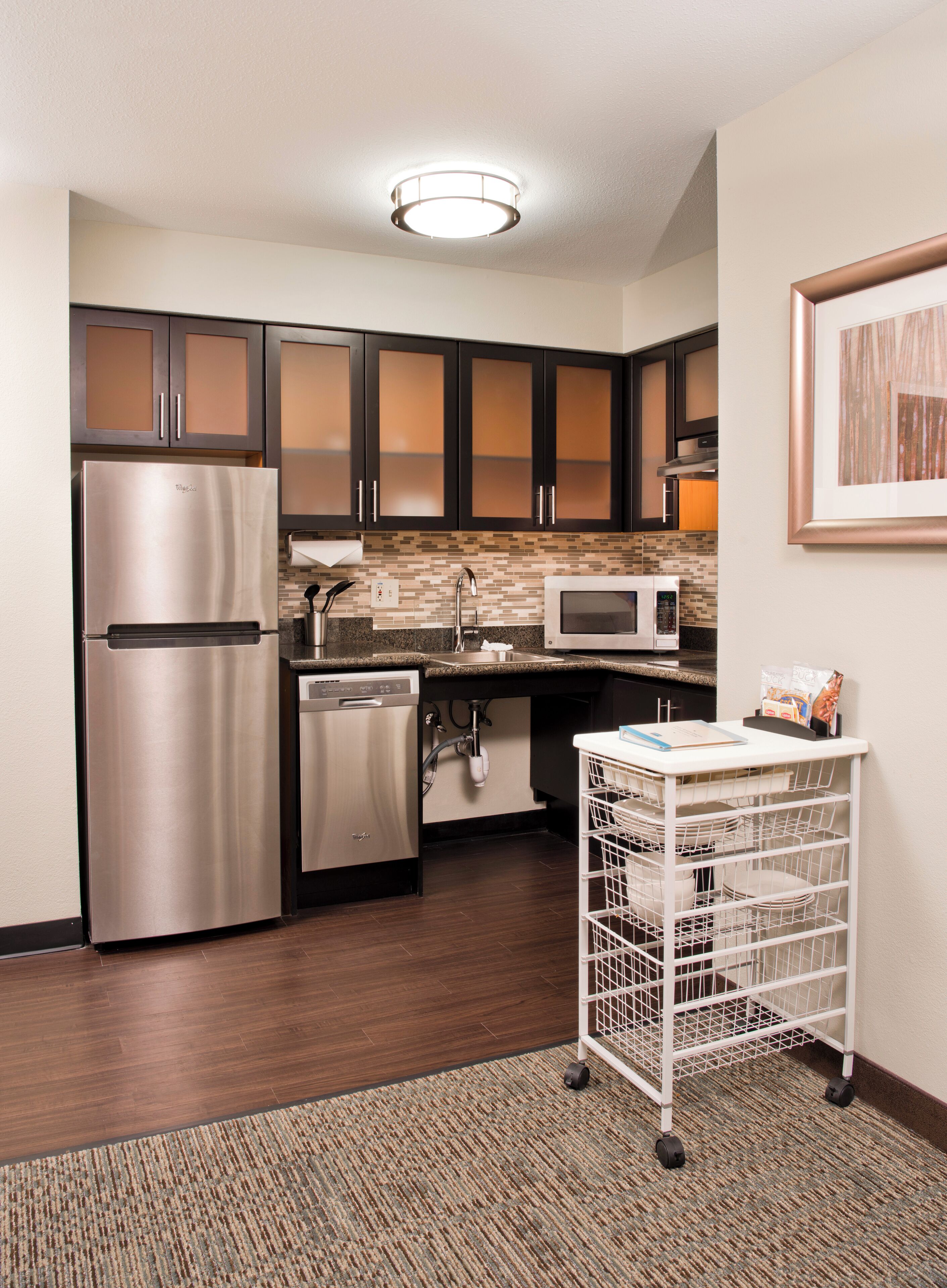 A compact kitchen with a stainless steel refrigerator, a dishwasher, a microwave, and a white cart with dishes.