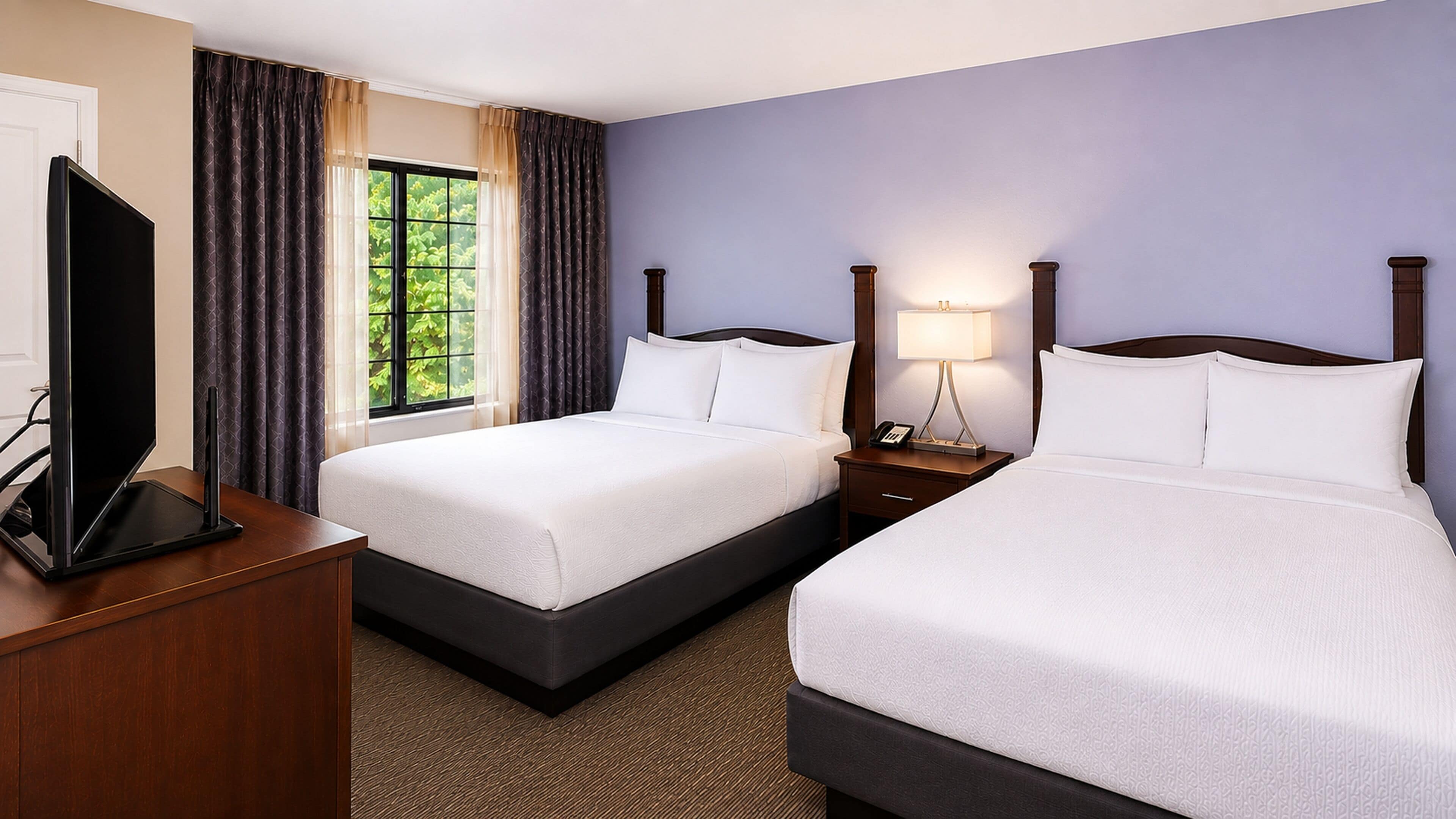 A hotel room with two beds, a desk with a flat-screen TV, a window with curtains, and a nightstand with a lamp.