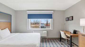 A hotel room with a bed, a desk, a chair, a radiator, a window with a view of the outside, and a picture on the wall.