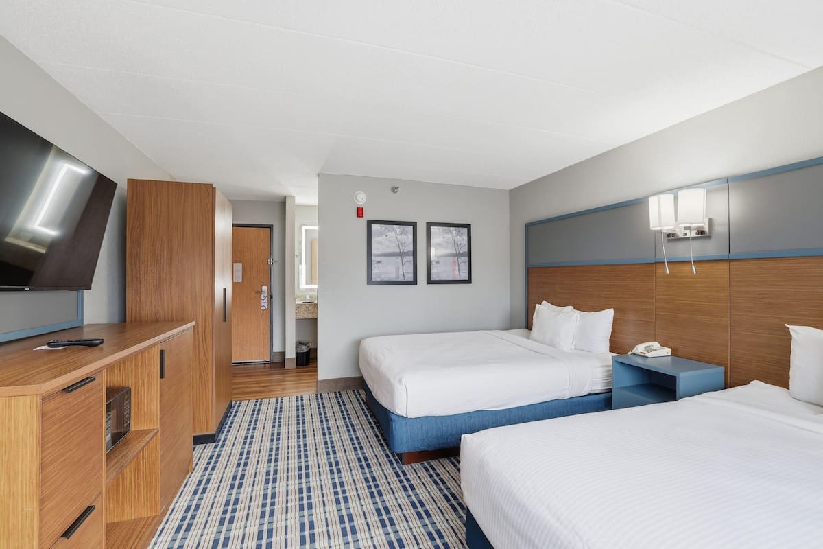Une chambre d’hôtel avec un grand lit, un plus petit lit, un bureau avec une télévision, et une salle de bain visible au fond.