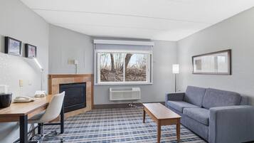 A hotel room with a fireplace, a sofa, a desk with a chair, a window with a view, and a patterned carpet.