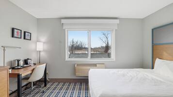 A hotel room with a bed, desk, chair, and a window with a view of a parking lot.