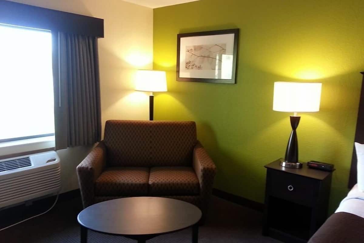 Une chambre d’hôtel avec un fauteuil brun, une petite table ronde, une table de chevet avec une lampe et une fenêtre avec des rideaux.