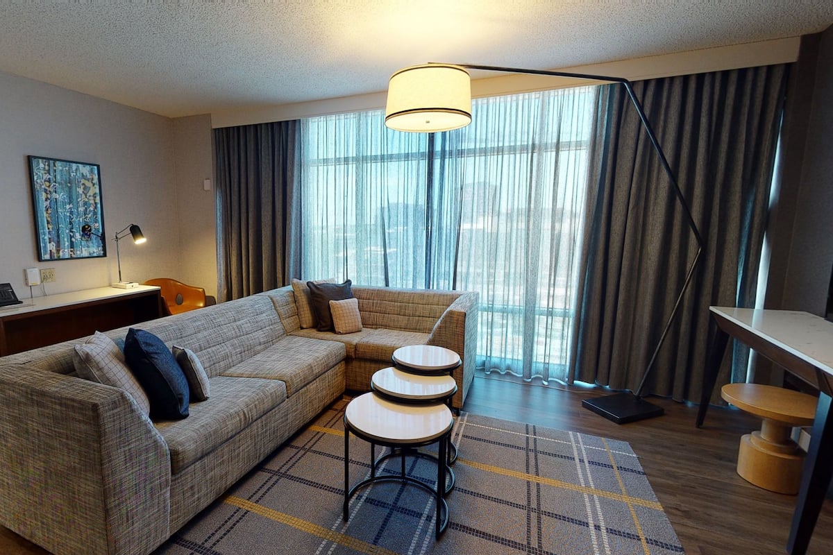 A modern hotel room with a sectional sofa, a small round table, a desk, and a large window with curtains.