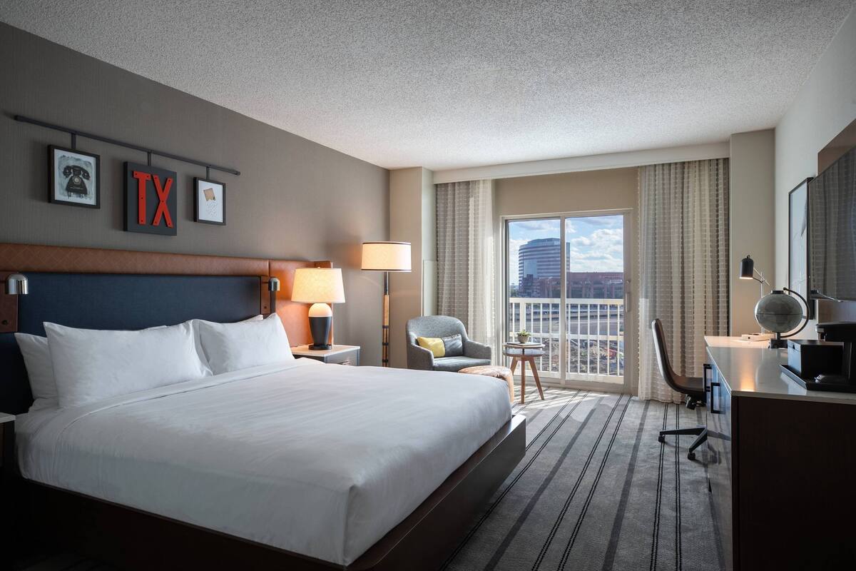 A hotel room with a large bed, a desk with a computer, a chair, a lamp, and a view of the city through the window.