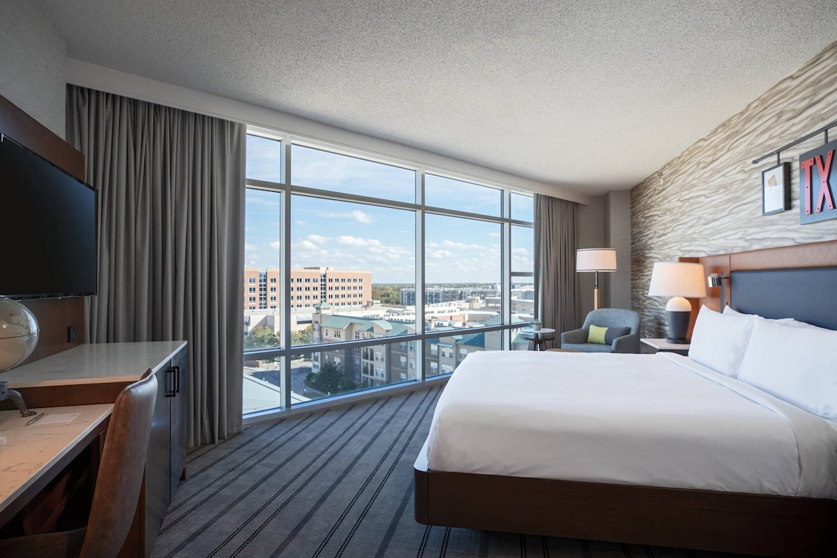 A hotel room with a large window, a bed, a desk, a chair, and a TV.