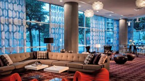 Un hall d’hôtel moderne avec un canapé d’angle, des fauteuils rouges, une table basse et de grandes fenêtres ornées de rideaux à motifs.