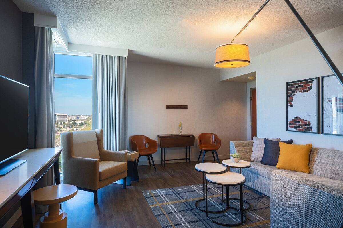 A modern hotel room with a large window, a sofa, a dining table, and a TV.
